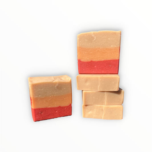 Five rectangular bars of layered handmade soap on a plain white background, with two bars standing upright and three stacked horizontally. The soap has a warm beige top layer, a peach-orange middle layer, and a red bottom layer.